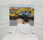 A couple sitting on a bed viewing out their window -- a perfect view of a backhoe where there ocean view once was